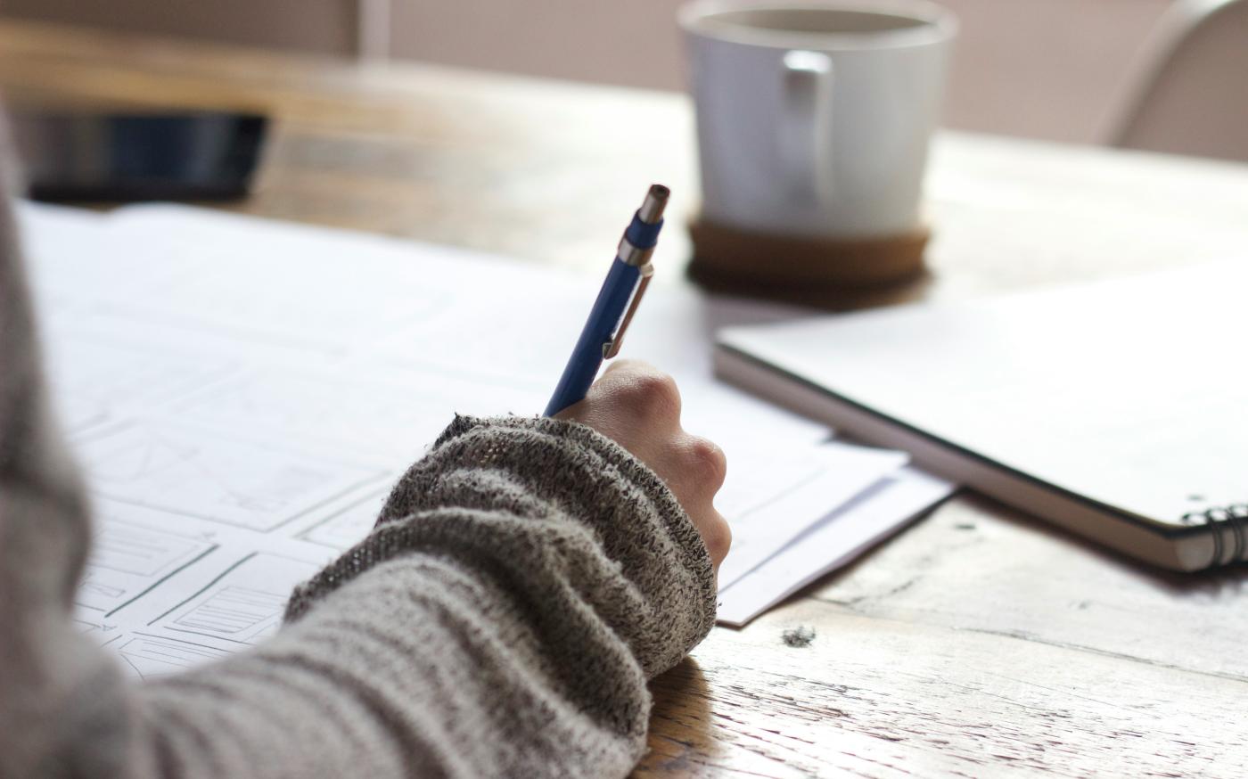 person writing on brown wooden table near white ceramic mug by Unseen Studio courtesy of Unsplash.